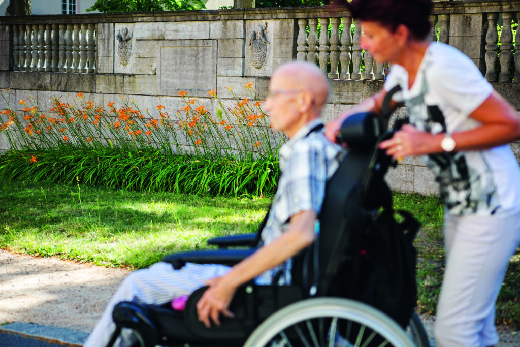 Palliative Care Weiterbildung. Pflegerin schiebt alten Mann in Rollstuhl vor grünem Gras und einer Mauer.