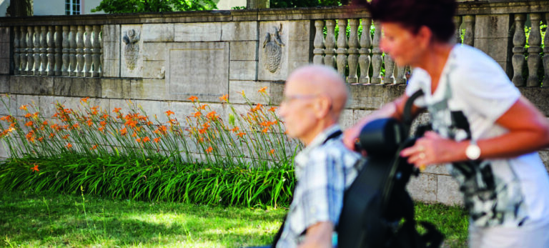 Palliative Care Weiterbildung. Pflegerin schiebt alten Mann in Rollstuhl vor grünem Gras und einer Mauer.